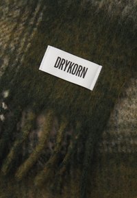 Mørkegrøn fuzzy tørklæde med frynseender og et hvidt mærke syet på, med fed sort tekst "DRYKORN".