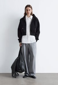 Schwarzer kurz geschnittener Hoodie mit silbernem Reißverschluss und Taschen, über einem weißen Hemd getragen. Graue Hosen mit weitem Bein, schwarze Tasche mit strukturiertem Rand.