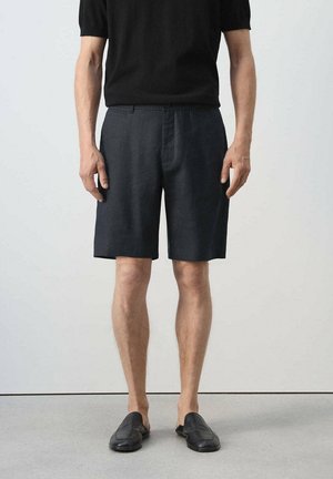 Man draagt donkere knielange shorts, zwart T-shirt met korte mouwen en zwarte instapschoenen, staand op een lichte vloer tegen een effen muur.