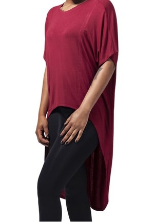 Bordowa oversize koszulka z krótkim rękawem i asymetrycznym dołem, wykonana z miękkiego materiału, w zestawieniu z czarnymi legginsami. Gładka faktura, luźny krój.