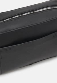 Sac en cuir noir avec une fermeture éclair en métal argenté visible et une poche avant ouverte, montrant une texture lisse et des détails de couture soignés.
