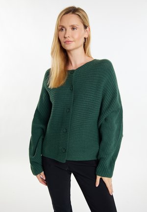 Zöld bordázott kardigán kerek nyakkal és oversized ujjakban, gombos elejével és struktúrált anyaggal. Fekete nadrág felett viselve.