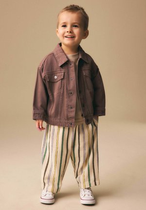 Copil mic zâmbind, stând în picioare, purtând o jachetă maro, o cămașă bej, pantaloni largi cu dungi și adidași albi, pe un fundal bej.