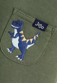 Grüner Stoff mit einem gestickten blauen Dinosaurierdesign und einem blauen Etikett mit der Aufschrift "Jojo" in der Nähe einer Tasche. Der Dinosaurier hat gelbe Akzente.