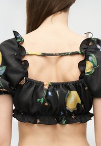 Οπίσθια όψη ενός ατόμου που φοράει μαύρο φλοράλ μίνι τοπ με φουσκωτά μανίκια και βολάν με ελαστικές ταινίες, αποκαλύπτοντας την άνω πλάτη και τους ώμους.