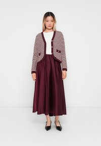 Γυναίκα φοράει λευκό μπλουζάκι, ριγέ μπουφάν μπορντό και λευκό, μακρύ μπορντό φούστα και μαύρες μυτερές γόβες, στέκεται μπροστά σε έναν λευκό φόντο.