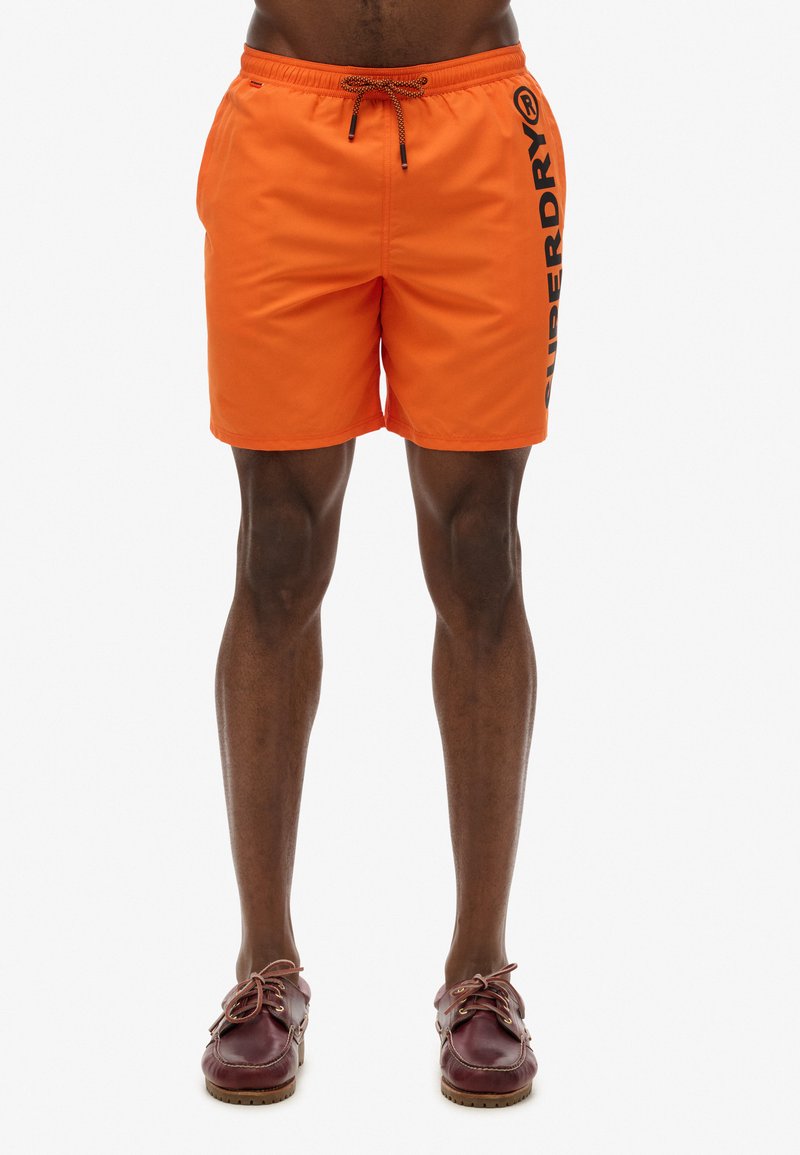 Shorts de bain orange vif avec une taille élastique, un cordon de serrage et un grand logo noir "SUPERDRY" sur le côté. Texture lisse.