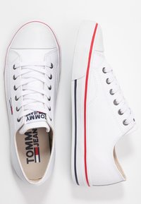 Białe płócienne trampki z okrągłym noskiem, białymi sznurówkami i gumową podeszwą, na której znajdują się czerwone i granatowe paski po bokach. Logo widoczne na języku.