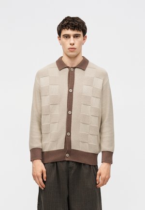 Ung mand iført beige og brun ternet strikket cardigan med knapper og mørke vidde bukser, står mod en ensfarvet hvid baggrund.