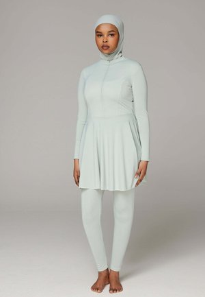 Femme portant un maillot de bain modeste bleu clair couvrant entièrement, avec manches longues, jupe, leggings et hijab de bain assorti, debout pieds nus sur un fond uni.