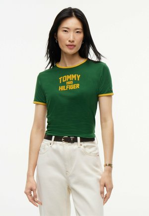 Donna con capelli neri che indossa una maglietta verde Tommy Hilfiger con rifiniture gialle e pantaloni chiari, in piedi davanti a uno sfondo bianco.