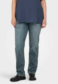 Plave traperice ravnog kroja, s izblijedjelim pranjem i glatkom teksturom, uparene s crnim sjajnim cipelama.