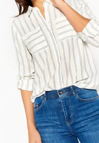 Vrouw in een witte overhemd met zwarte strepen en opgerolde mouwen, gecombineerd met blauwe denim jeans.