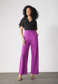 Blusa nera a scollo a V con maniche trasparenti, abbinata a pantaloni viola a gamba larga e alta vita, e sandali neri con cinturini. Texture lisce.