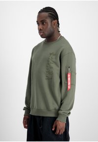 Felpa verde oliva con logo in rilievo "ALPHA INDUSTRIES", collo a giro, e tasca con zip sulla manica con etichetta rossa.