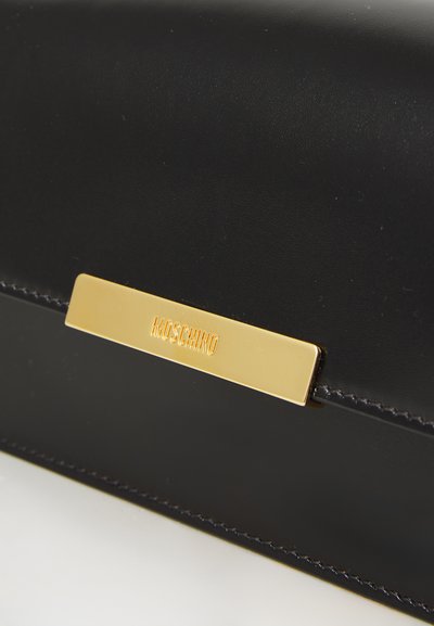 Crna kožna torbica sa glatkom teksturom, sa zlatnom metalnom pločicom ugraviranom s natpisom "MOSCHINO" i kontrastnim šavovima.