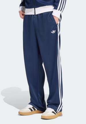 Person trägt dunkelblaue Adidas-Trainingshose mit weißen Streifen und Logo, kombiniert mit weißen Sneakers mit schwarzen Streifen und Gummisohlen.