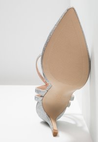 Chaussure à talons aiguilles argentée scintillante avec un bout pointu, un talon fin et deux brides de cheville. Semelle beige lisse avec texture nervurée et marquage.