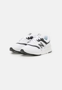 Pár bílých a černých sportovních bot New Balance s tkaničkami, bočním logem a silnými gumovými podrážkami na bílém pozadí.