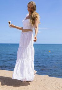 Top corto bianco in pizzo con spalline, abbinato a una gonna maxi bianca a più strati, entrambi con motivi intricati, indossati con sandali.