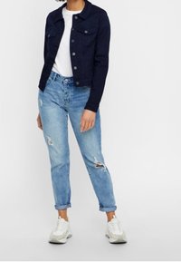 Giacca di jeans blu navy con due tasche sul petto, t-shirt bianca sotto, jeans chiari strappati e sneakers bianche con dettagli grigi.