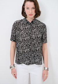 Blusa nera a maniche corte con motivo floreale bianco, chiusura frontale con bottoni e orlo arrotondato. Bracciale in argento al polso. Indossata con pantaloni bianchi.