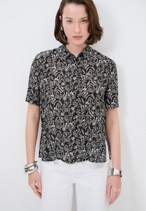 Blusa nera a maniche corte con motivo floreale bianco, chiusura frontale con bottoni e orlo arrotondato. Bracciale in argento al polso. Indossata con pantaloni bianchi.