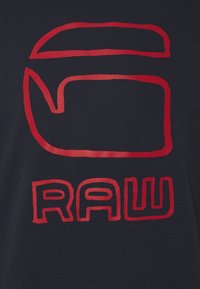 Maglietta blu navy con un audace logo grafico rosso, che comprende una lettera stilizzata e la parola "RAW", con una texture del tessuto liscia.