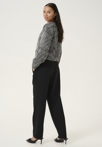 Kobieta o długich ciemnych włosach, ubrana w szary sweter o fakturze, czarne szerokie spodnie oraz czarne mulesy na obcasie, stojąca z rękami w kieszeniach.