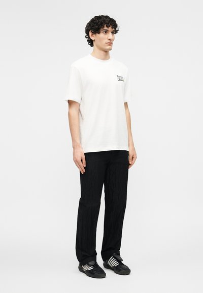 Νεαρός με σγουρά μαλλιά που φορεί λευκό T-shirt με σχέδιο, μαύρο παντελόνι με υφή και μαύρα αθλητικά παπούτσια Adidas με λευκές ρίγες.