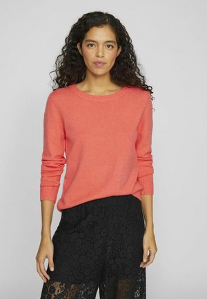 Pull en tricot corail avec un col rond et des manches longues, associé à un pantalon en dentelle noir avec des motifs floraux et des éléments transparents.