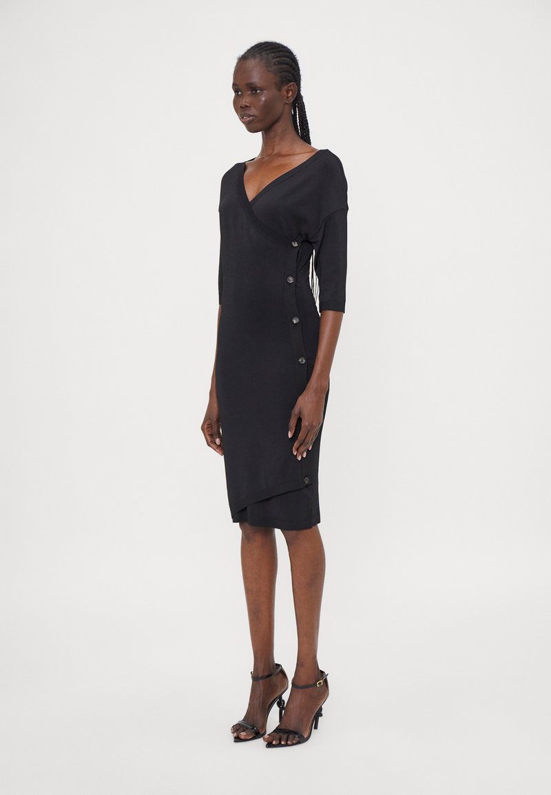 Schwarzes Wickelkleid mit tiefem V-Ausschnitt, dreiviertellangen Ärmeln und seitlichen Knopfdetails. Glatte Textur, knielang, getragen mit schwarzen Absätzen.