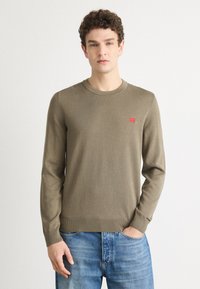 Olivengrønn strikkegenser med rund hals, ribbekanter på ermene og nederkant. Liten rød merkelapp på brystet. Kombinert med blå denimjeans.