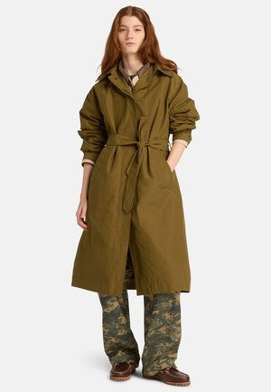 Olivgrüner Trenchcoat mit tailliertem Gürtel, langen Ärmeln und Kragen. Das Modell trägt camouflagegemusterte Hose und braune Schuhe.