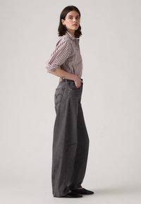 Jauna sieviete stāv profilā, valkājot sarkano un baltu svītraino krekli, brīvās kājas augsto jostasvietu melnos džinsos un melnās norādītājkurpēs.