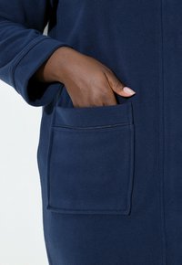 Tmavě modrý fleecový župan s našitou kapsou, měkkou texturou a rukávem s přímým střihem, s rukou umístěnou uvnitř kapsy.