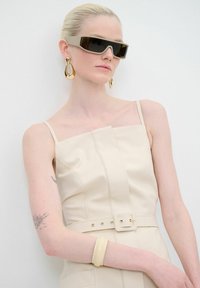 Robe en cuir beige avec un décolleté carré, une taille ceinturée et des fines bretelles. Des lunettes de soleil noires et des boucles d'oreilles en forme de goutte d'or complètent le look.