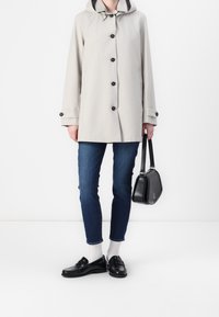 Persoon in een beige gewatteerde jas met capuchon, blauwe jeans, witte sokken, zwarte instappers, die een zwarte leren handtas vasthoudt tegen een witte achtergrond.