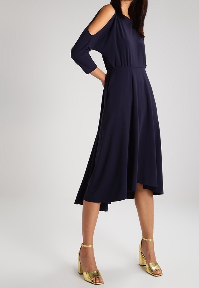 Femme portant une robe midi bleu marine avec épaules découpées et sandales à talons hauts dorées, debout devant un fond clair uni.