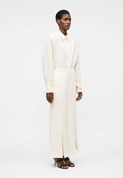 Γυναίκα που φοράει ένα εκρού υπερμεγέθη πουκάμισο με κουμπιά, μαζεμένο μέσα σε ψηλόμεσα παντελόνια με φαρδιά μπατζάκια και μπροστινές σχισμές, συνδυασμένα με μαύρα σανδάλια με τακούνι.