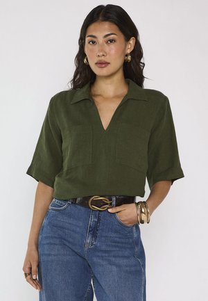 Femme portant une chemise à col vert olive, un jean bleu, des créoles dorées, des bracelets en or et une ceinture à boucle serpent, debout devant un fond uni.