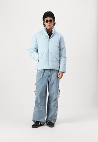 Light blue puffer jacket with a zip front, featuring a high collar and quilted design, paired with loose, light blue cargo jeans and black shoes.