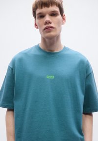 Τυρκουάζ κοντομάνικη μπλούζα από απαλό, υφασμάτινο ύφασμα με υφή. Διαθέτει ένα ξεχωριστό πράσινο λογότυπο "HUGO" στο μπροστινό μέρος, στο κέντρο κάτω από τη λαιμόκοψη.