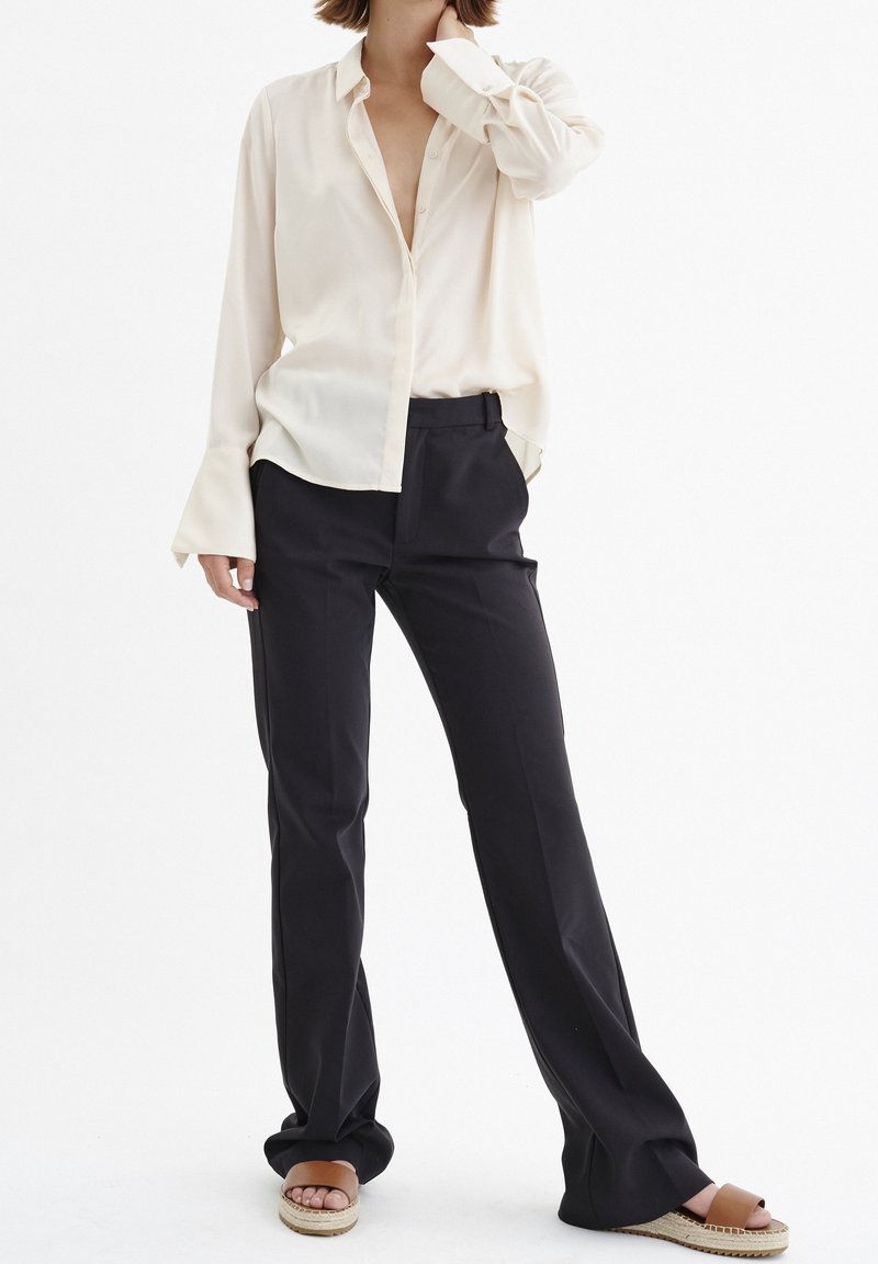 Femme portant une chemise ample crème à boutons, un pantalon noir à jambes larges et des sandales espadrilles marron clair sur fond blanc.