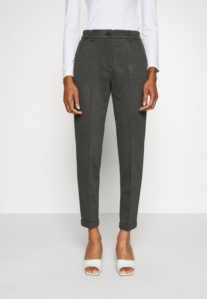 Pantalon à motif pied-de-poule gris avec une coupe fuselée, présentant un devant plat, des passants de ceinture et des ourlets roulés. Porté avec des chaussures blanches ouvertes.