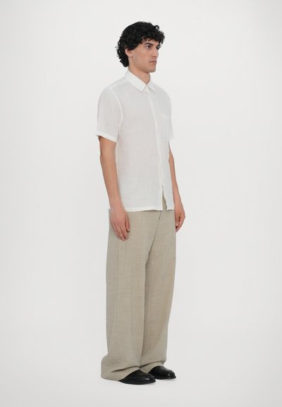 Camisa branca de manga curta com botão e bolso, combinada com calças largas bejes claras e sapatos pretos, de pé contra um fundo liso.