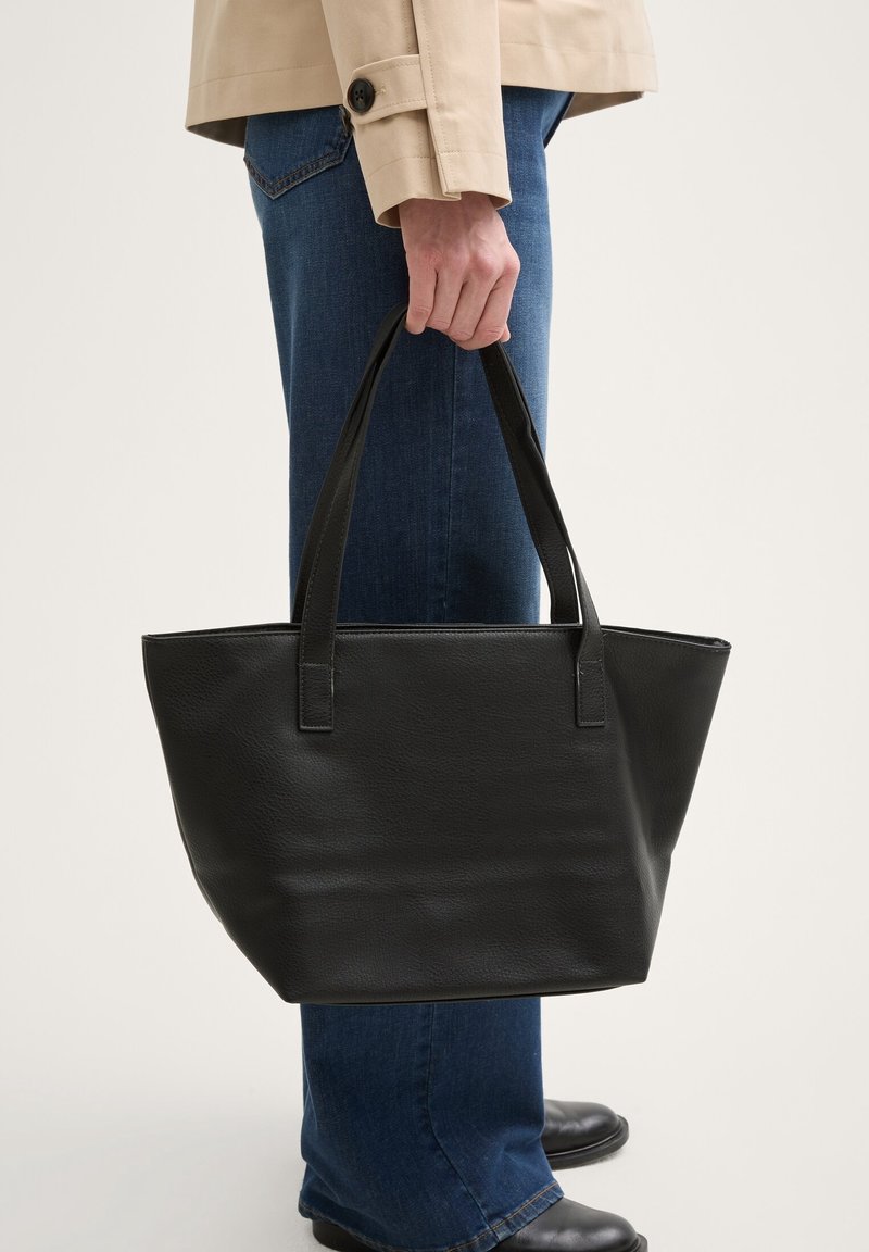 Personne portant un manteau beige et un jean bleu tient un grand sac cabas en cuir noir par ses anses à côté d'elle.