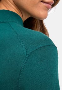 Pull en maille teal avec un col montant, un col côtelé et une texture lisse, présentant des détails de couture visibles sur l'épaule.