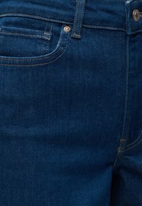 Gros plan sur un jean en denim bleu foncé montrant la poche avant, la boucle de ceinture, les coutures et les rivets en métal.