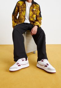 Abbigliamento sportivo da uomo: una giacca nera e gialla a fantasia, pantaloni neri larghi e scarpe bianche con dettagli bordeaux. Seduto su uno sgabello di metallo.